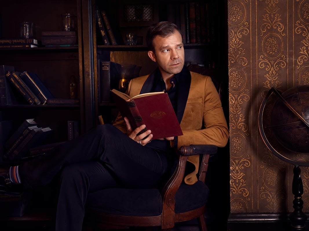 A wide shot of Critical Role main cast member Liam O'Brien. He's wearing a brown blazer and a black button-down shirt, and he's holding a red, leather-bound book.