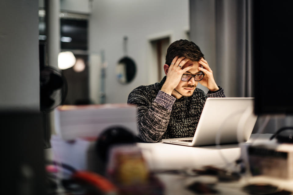 Young casual businessman wearing glasses is sitting in front of his notebook holding his head pondering over his work. Office equipment and another computer is in front of him.