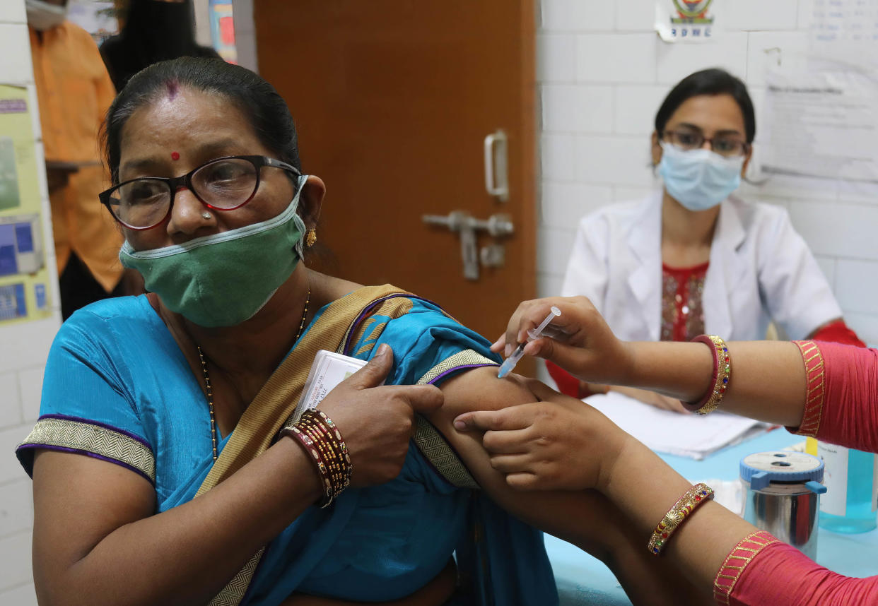 A woman in a blue sari wearing a green mask bares her arm for a shot in a medical office, with another health worker in white scrubs sitting at a desk behind her. 