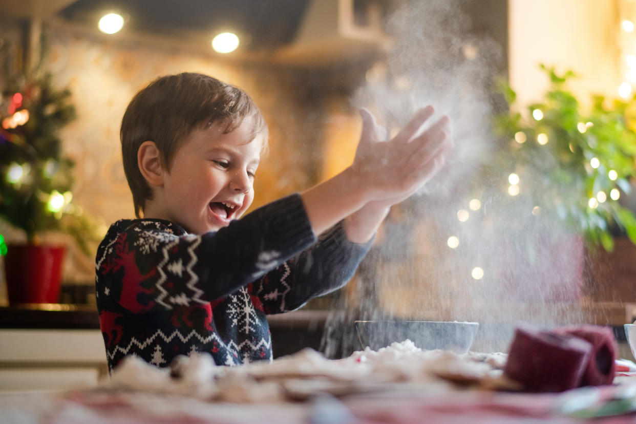 A child wearing a sweater decorated with reindeer, snowflakes and fir trees stands above a mixing bowl apparently clapping their hands as powder erupts around them.