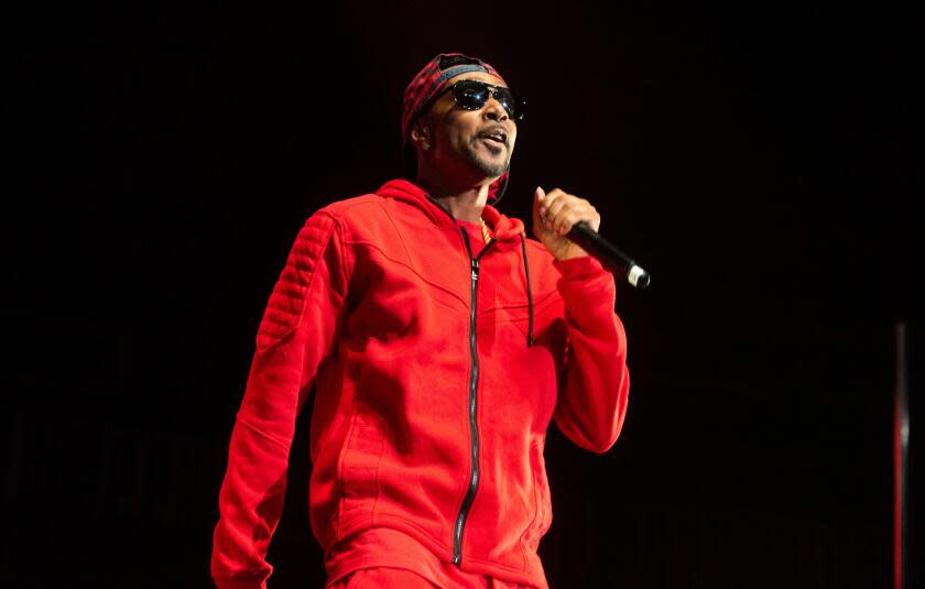 Krayzie Bone stands on a darkened stage in a red tracksuit, dark glasses and backward cap and raps into a microphone