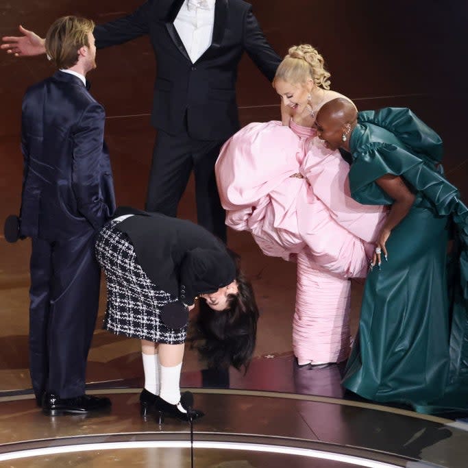 Group of celebrities at an event with one bending down and the others reacting to a moment on stage