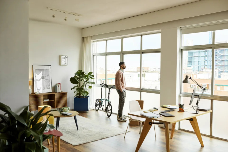 Full length of young man with hands in pockets standing in living room. Man is looking through window at home. He is wearing casuals.