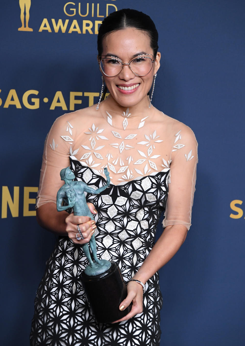 Ali Wong posing with SAG award, wearing a patterned dress and clear glasses, smiling at the camera