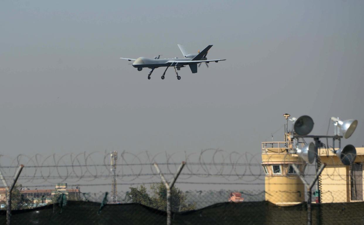 A drone aircraft is aloft, wheels down, above a razor-wire fence and yellow control tower.