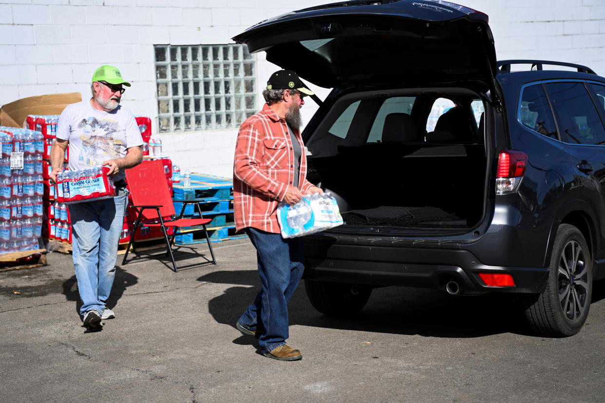 Larry Culler, with a white beard and a fluorescent green baseball cap and Ed Byard, in beard and plaid shirt, load batches of water into the back of an SUV.