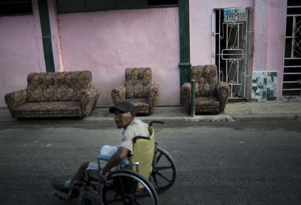 In this April 12, 2014 photo, a man in a wheelchair passes next to a living room set sitting on the steet to dry, next to a poster of a house that reads "Swap Agreement" in Havana, Cuba. Around Havana Cubans can be seen taking advantage of the materials now available as they add second stories to their homes, enclose balconies to turn them into an extra room or throw on a fresh coat of paint. (AP Photo/Ramon Espinosa)