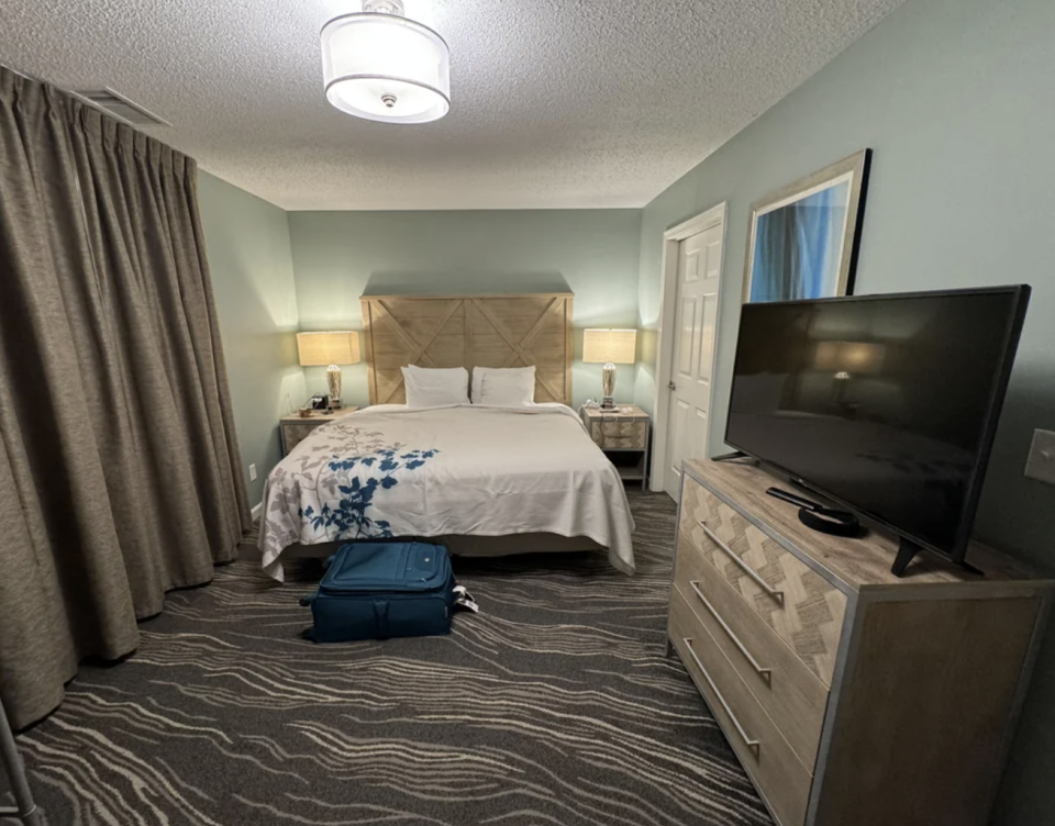 Hotel room with a bed, side tables, TV, dresser, and a blue suitcase on the floor