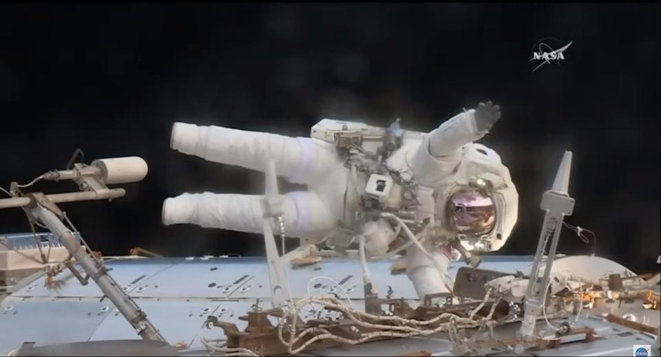 An astronaut in a spacesuit floats above a module.  He holds it with one hand and waves with the other.  is on its side from the angle the photo was taken