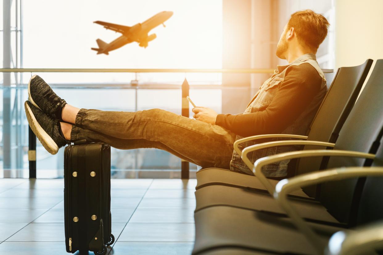 A man in an airport, sitting with his legs propped up on a carry-on, watching a plane take off. 