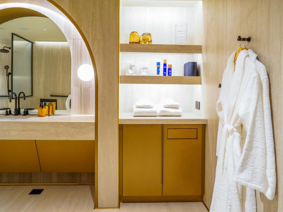 Inside a bathroom with large mirrors and gold accents. On the left are two hanging white robes and a shelf with toiletries