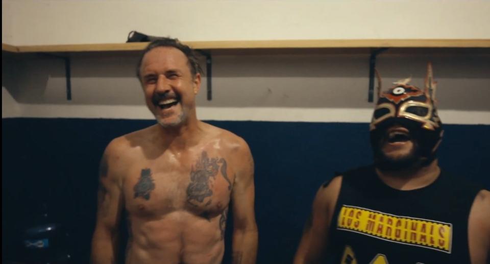 David Arquette laughs alongside a luchador in a locker room