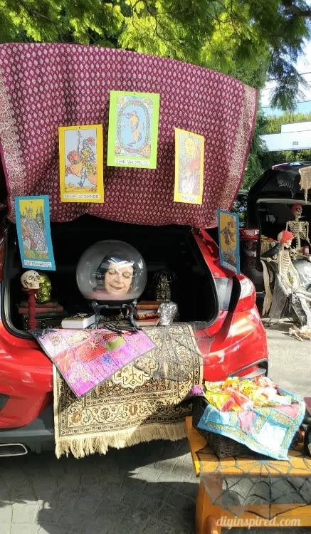 open trunk of car with a large crystal ball with head inside of it, skulls stacked on books, an oriental carpet hanging out of it, and a table in front of the trunk with a spiderweb on it and a basket full of candy on it