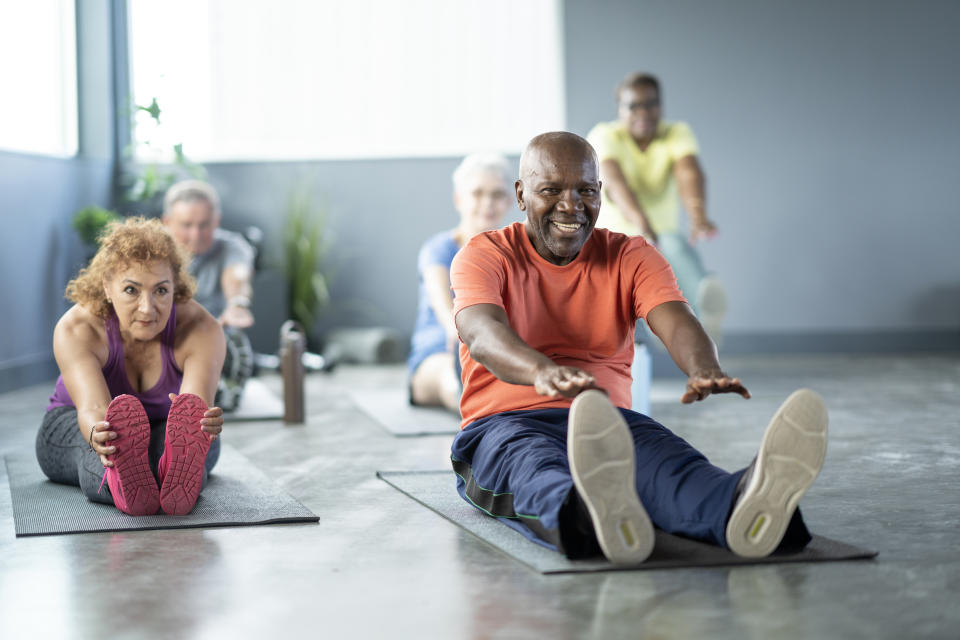 A group of people in a yoga class, sitting on mats and reaching forward in a stretch. They are focused and smiling, engaged in their exercise routine