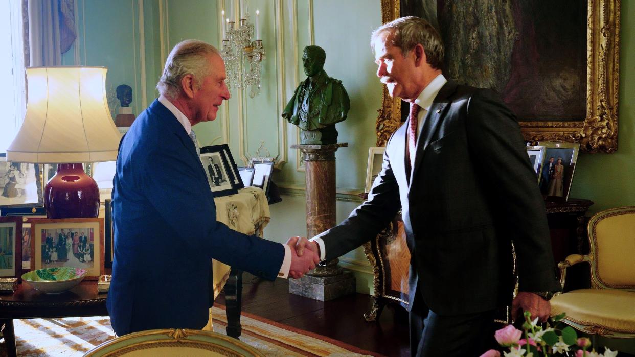  king charles iii shaking hands with chris hadfield in a palace room surrounded by photos, chairs, tables. a statue is on a pillar between them 