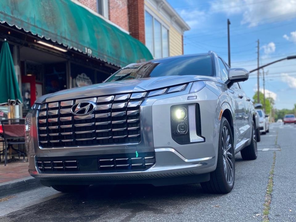 A silver Hyundai Palisade SUV with its headlights on parked on the street in front of a restaurant.