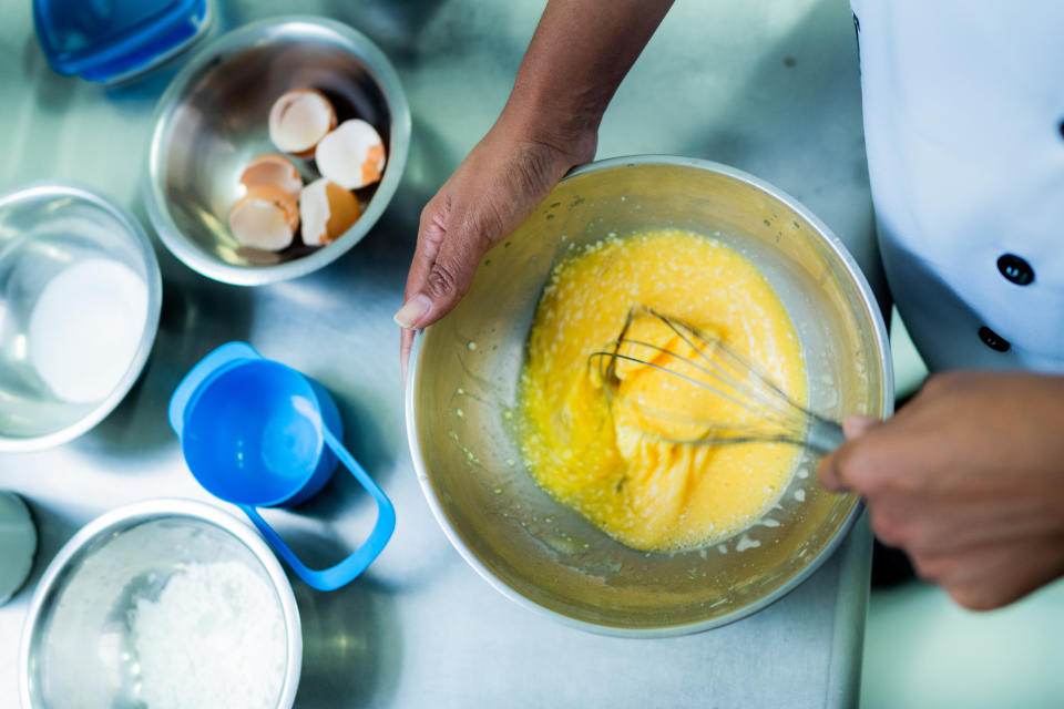 Person whisking eggs in a bowl, surrounded by bowls containing eggshells, flour, and a blue measuring cup on a kitchen countertop