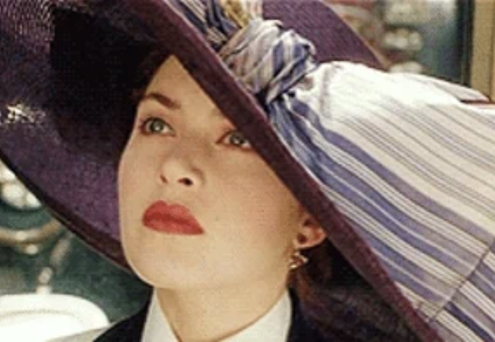 Kate Winslet in Titanic, wearing an elegant hat with a striped ribbon and looking upward pensively