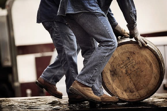 Two people in jeans pushing an oak barrel along the floor.