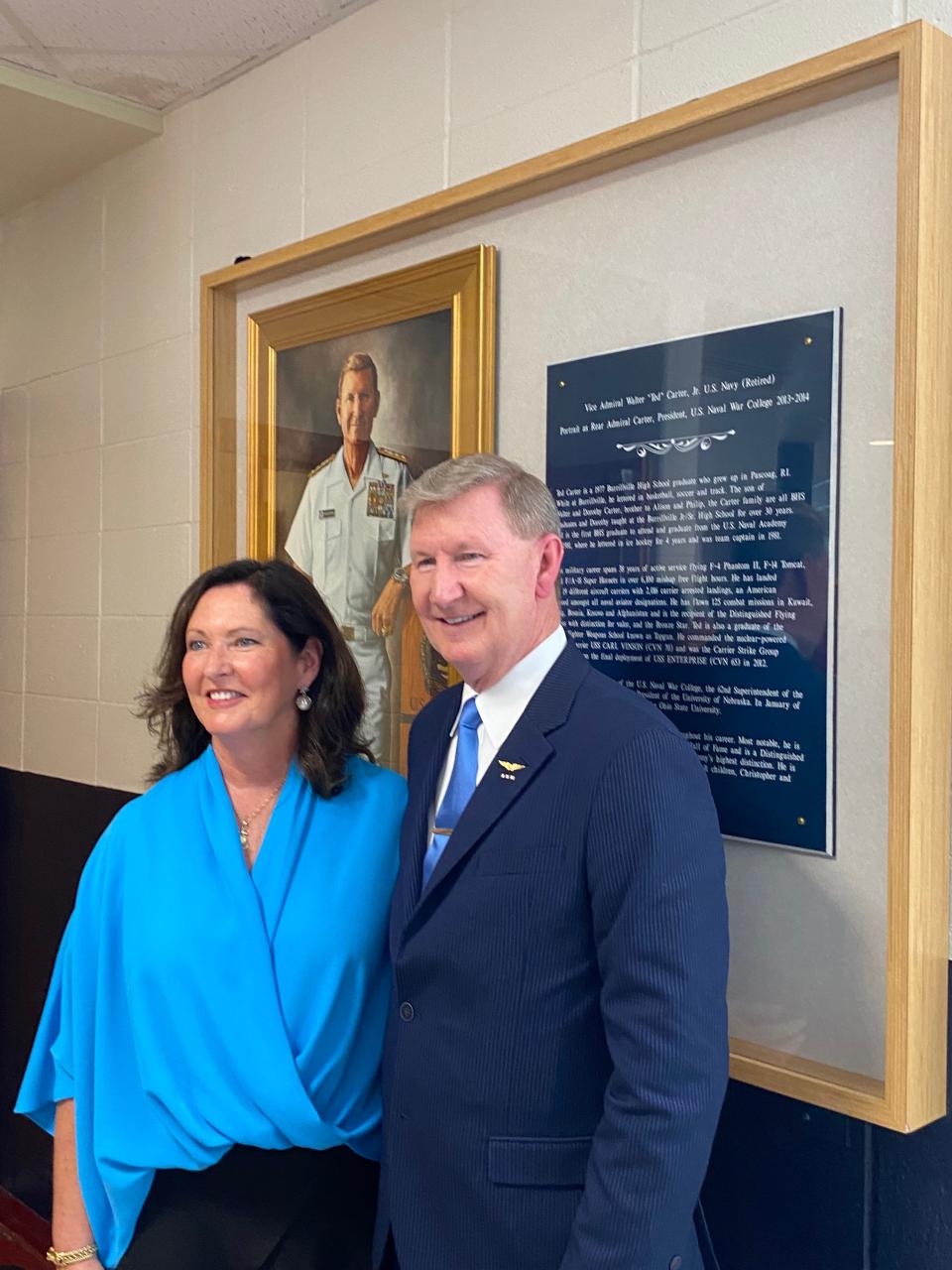 Retired Navy Vice Adm. Walter "Ted" Carter and his wife, Lynda, stand before the just-unveiled shadow box at the doors of the gymnasium renamed in his honor in Burrillville. The portrait in the background depicts him as president of the Naval War College in Newport.