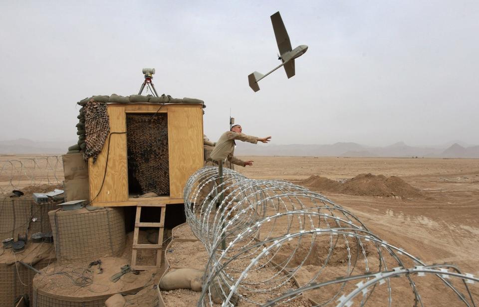 A Marine launching a drone from behind barbed wire, next to a hut with a ladder going up to it.