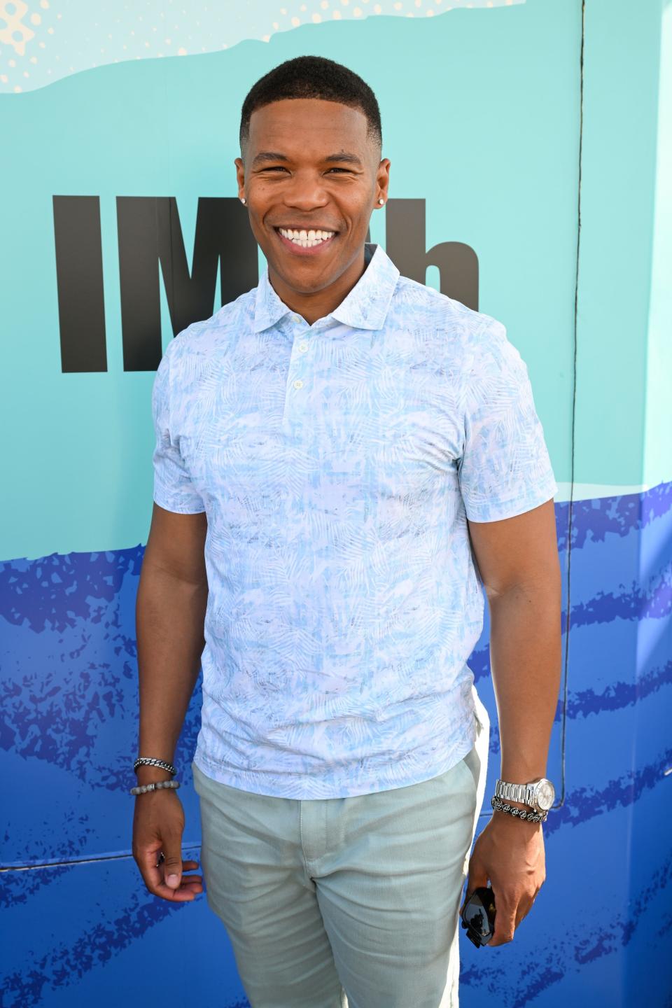 A person is smiling, wearing a patterned short-sleeve shirt and light pants, standing in front of an IMDb backdrop at an event