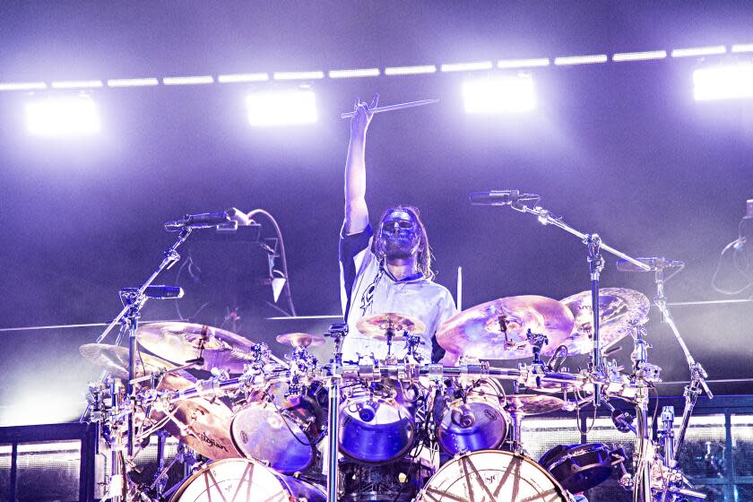 A man wearing a mask sitting behind a drum kit on a stage holding up his drumstick in his right hand.