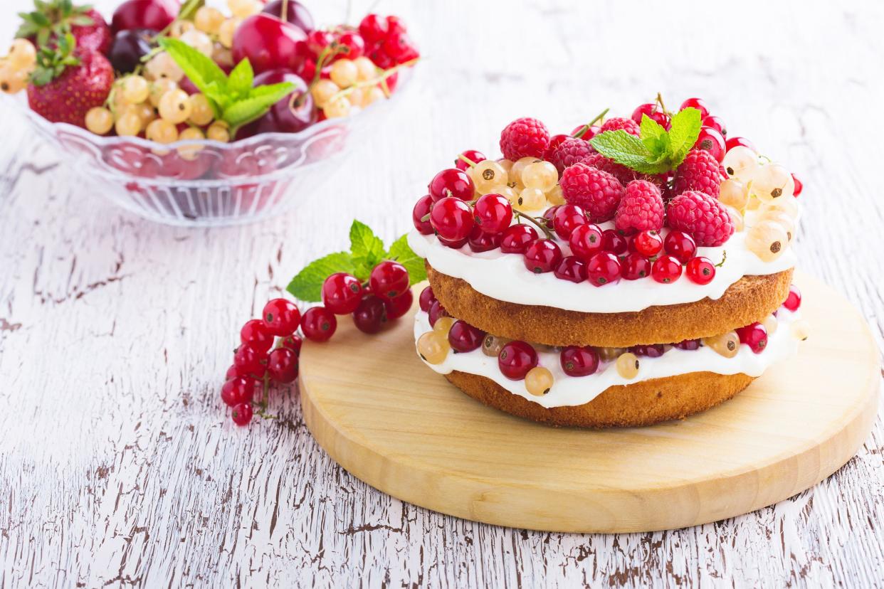 Coconut cream & fruit-topped vanilla cake on a round wooden cutting board with a bowl of berries in the background