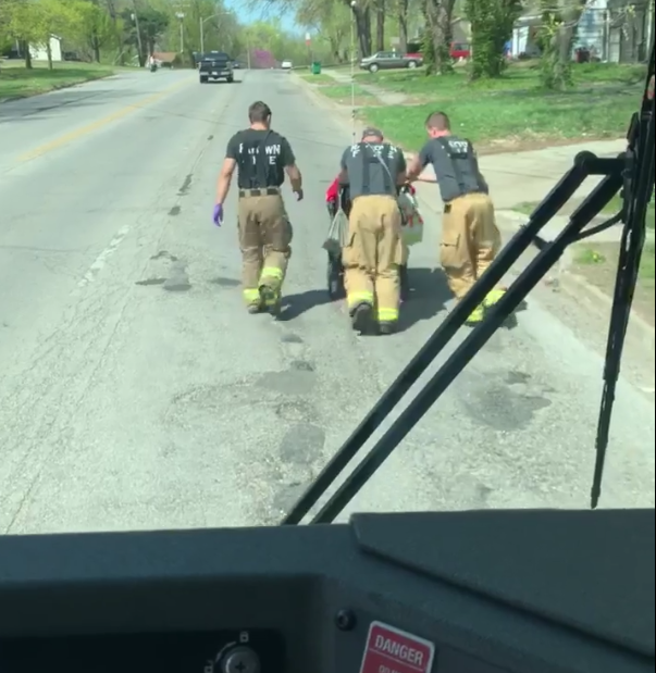 Drei Feuerwehrmänner schieben einen Rollstuhlfahrer eine Anhöhe hinauf. Das Video der guten Tat ist aus dem Fahrerhaus des Einsatzwagens aufgenommen, der Scheibenwischer steht halb im Bild. Foto: Screenshot / Facebook / Raytown Fire Protection District