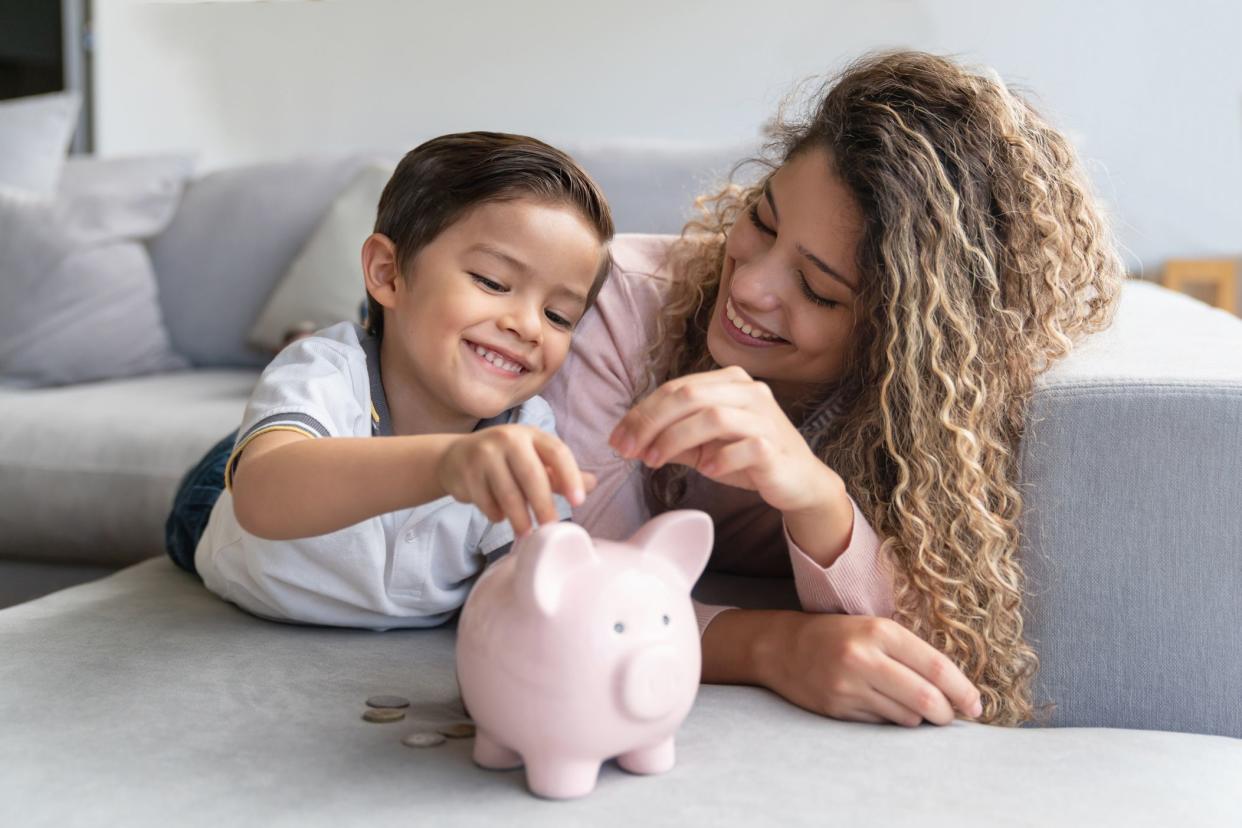 Son on left, mother on right, both are smiling and putting coins in a piggybank while laying on a grey couch, facing towards the camera with rest of 'L'-shaped couch and grey wall of living room in the background