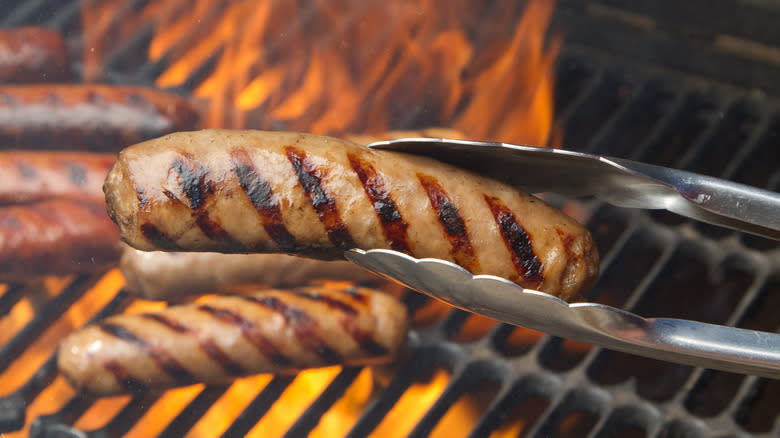 A bratwurst in a pair of tongs over a flaming grill