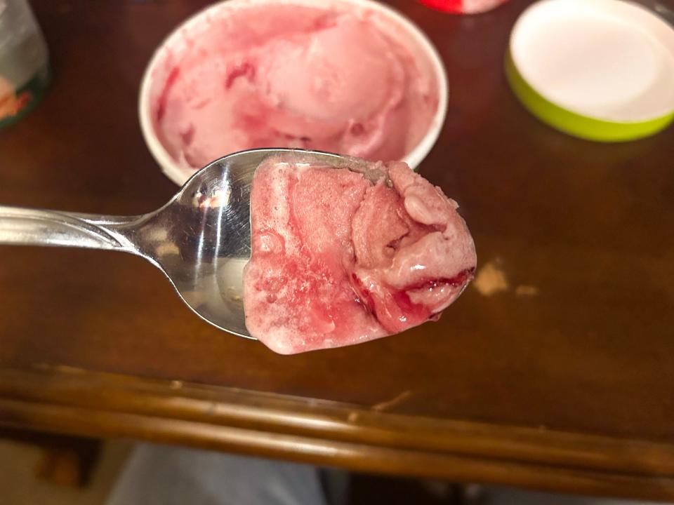 A metal spoonful of pink frozen dessert with a darker purple swirl in it with a carton of dessert in the background