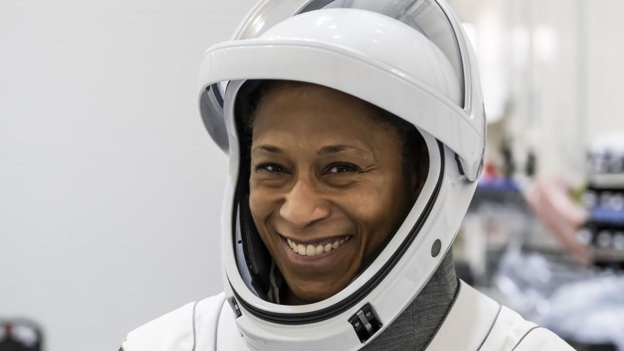  Woman smiling in a spacesuit with lab equipment far in the background. 