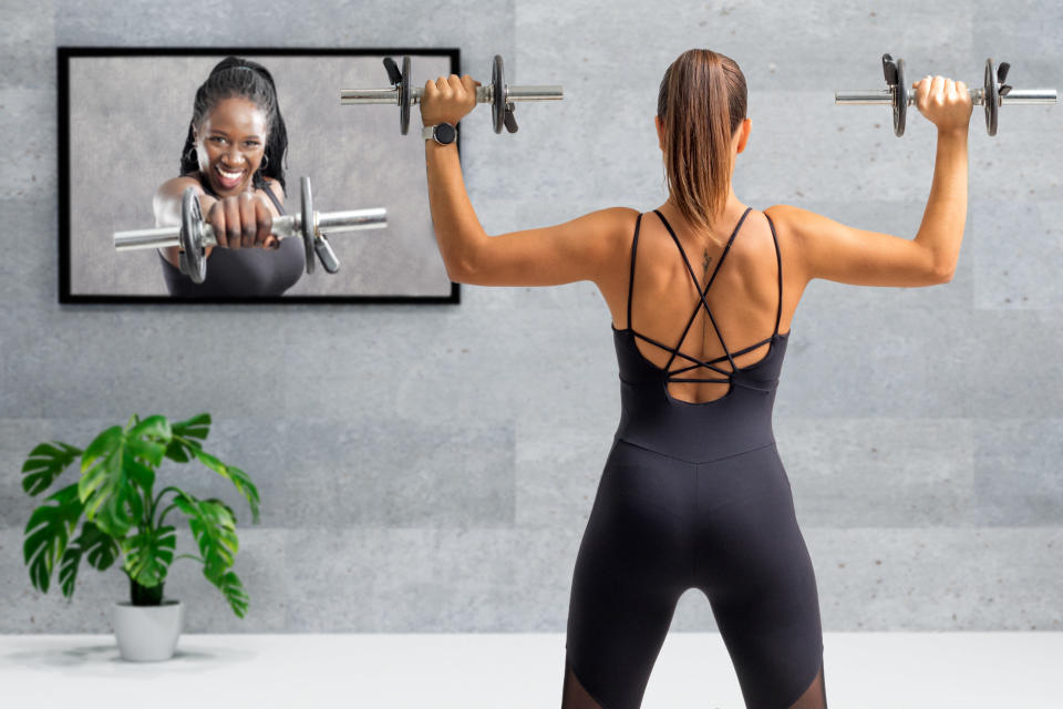 Rear view of young woman working out with dumbbells at home while watching a female trainer on a screen holding a weight.