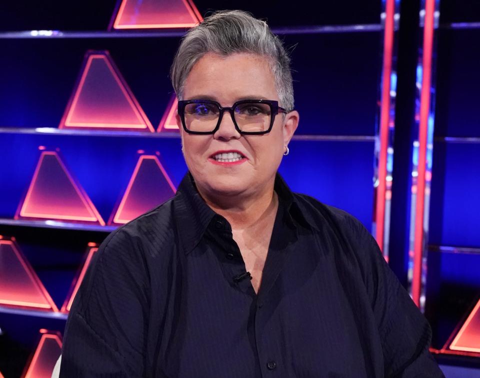 Rosie wears a black button down while smiling on the set of a game show