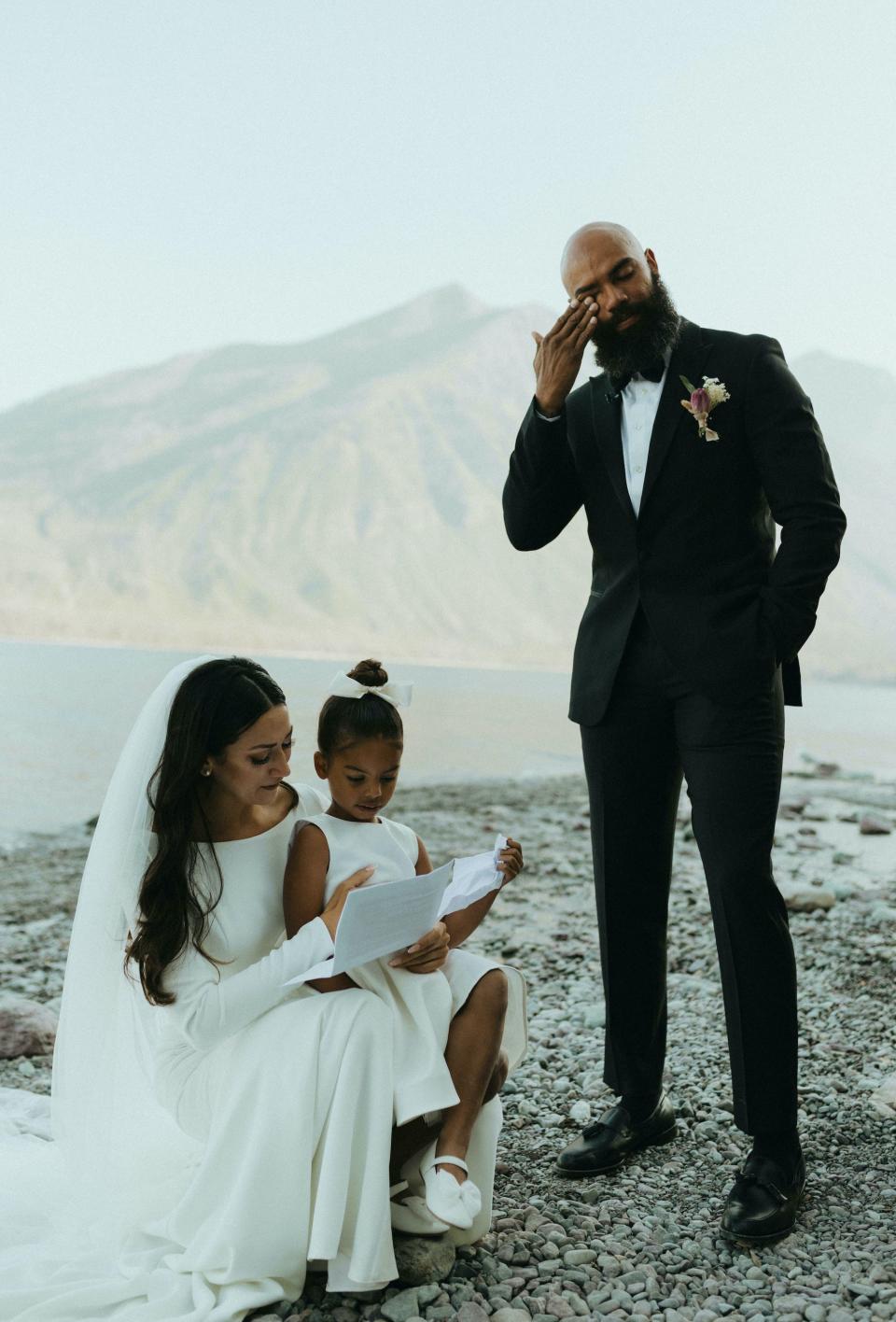 A groom wipes away a tear as a bride and a flower girl read a letter for him crouched on the ground.