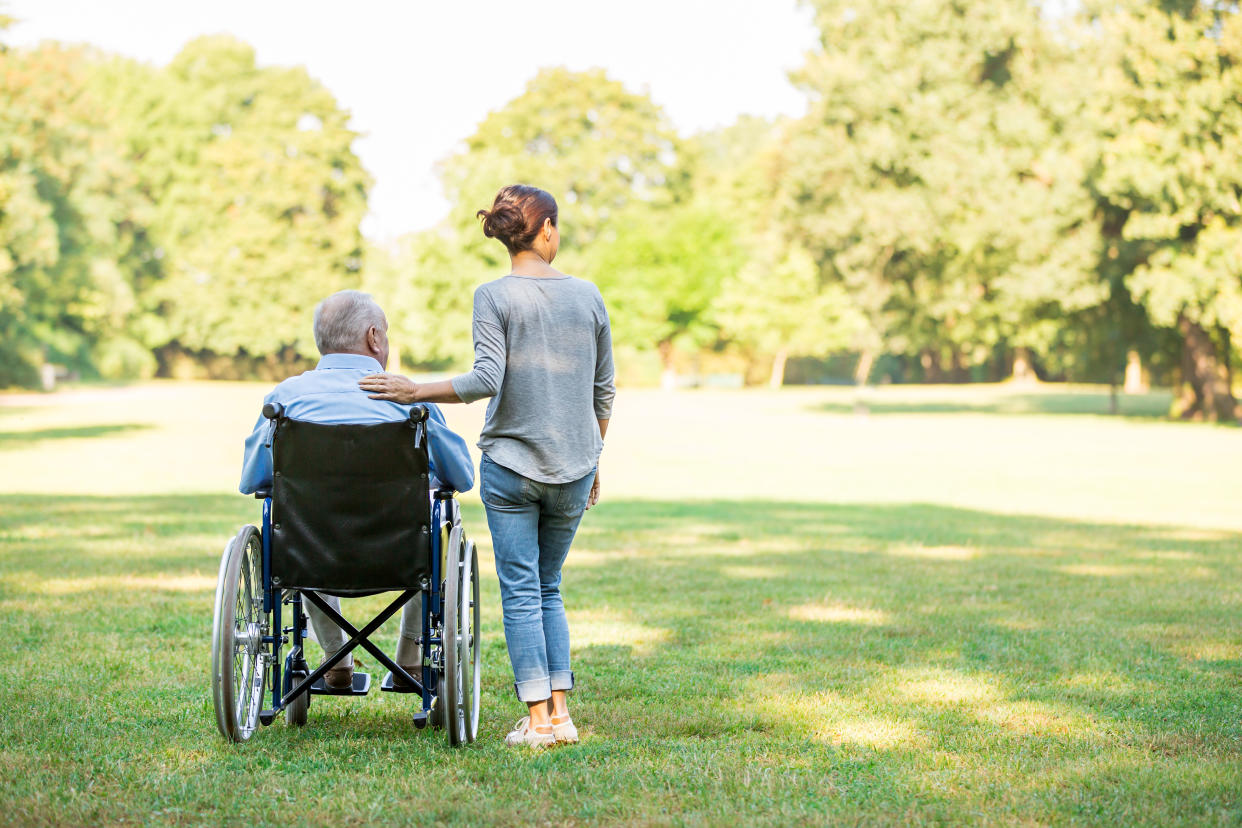 Senior man sitting in a wheelchair on a lawn, with caregiver standing next to him.