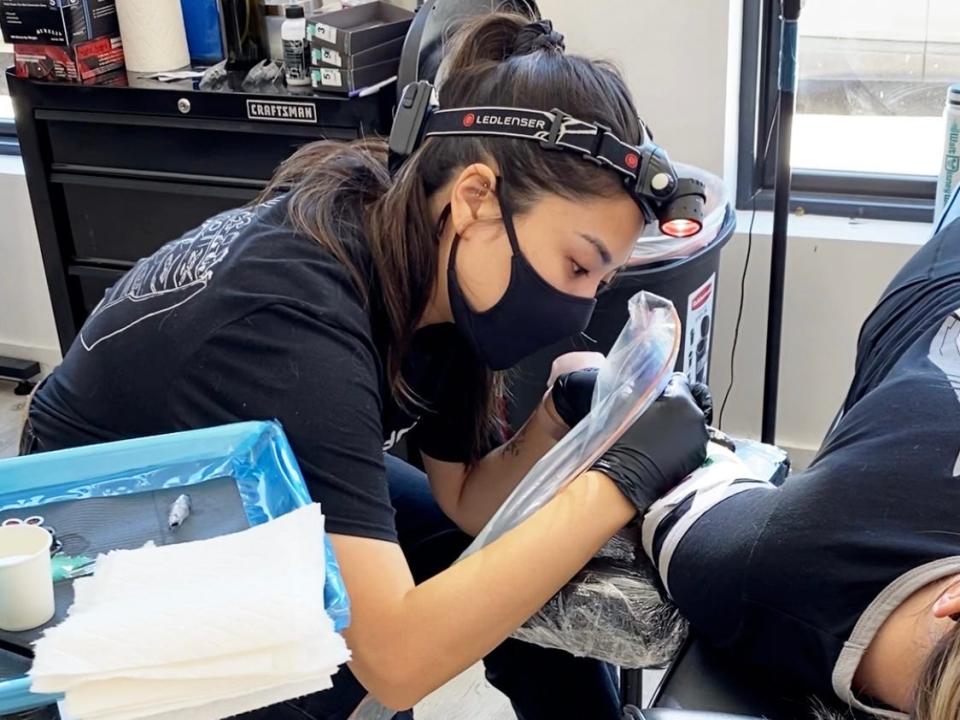 The writer wearing black and tattooing someone's arm in a tattoo shop