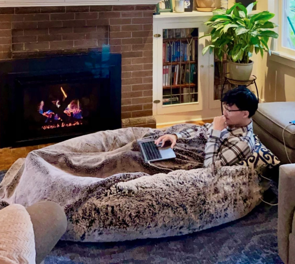 A man sits snuggled in a Plufl cushion bed working on his laptop in a loungeroom with a warm fire.