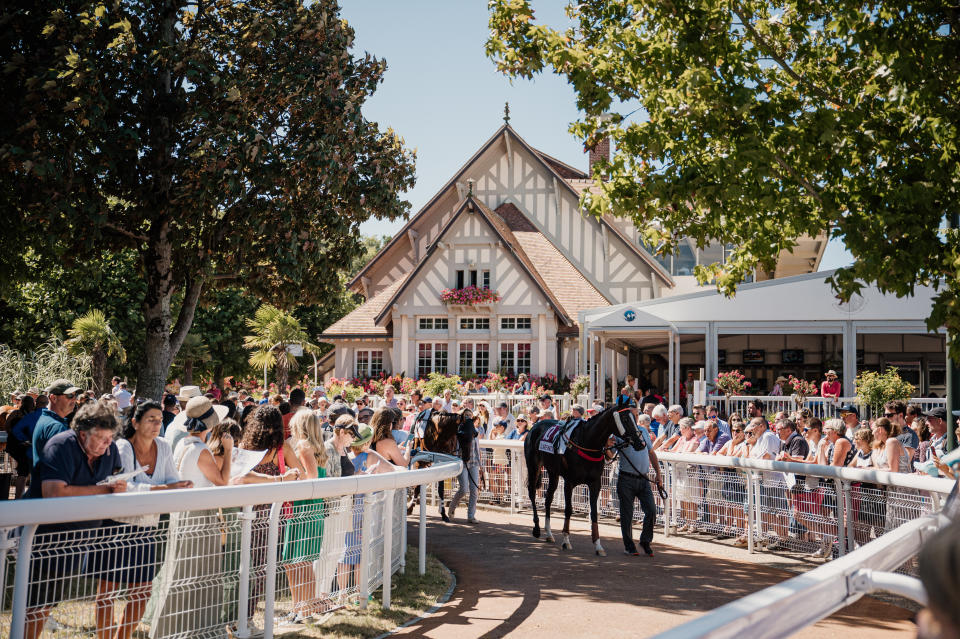 An image of Clairfontaine racecourse with attendees on both sides of a course with two horses being walked down it.