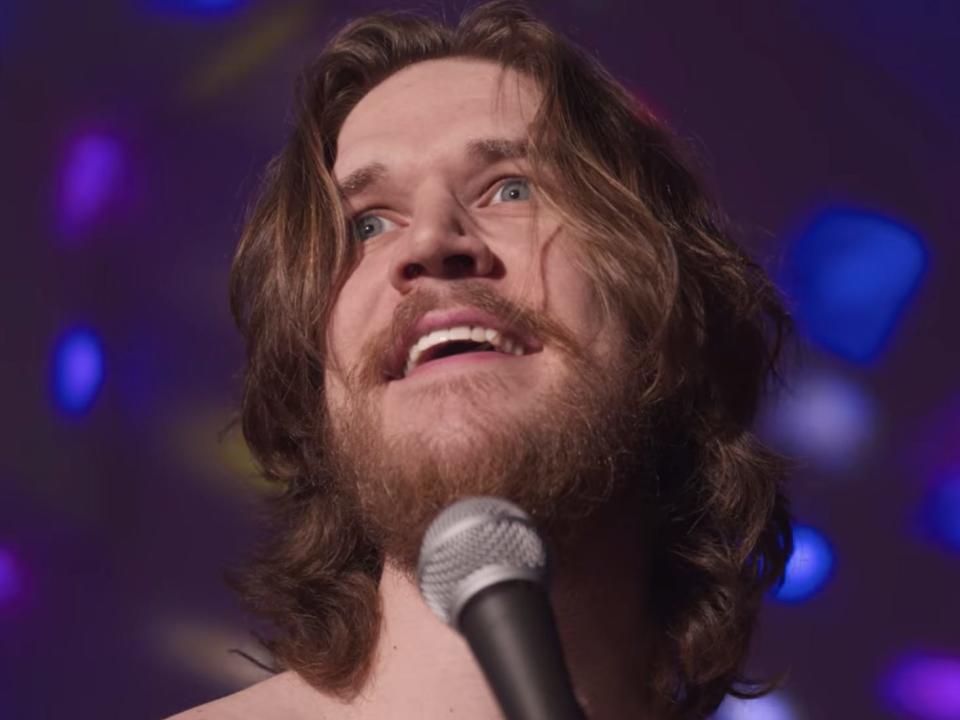 A close up of a man (Bo Burnham) singing into a microphone with dotted lights behind him.