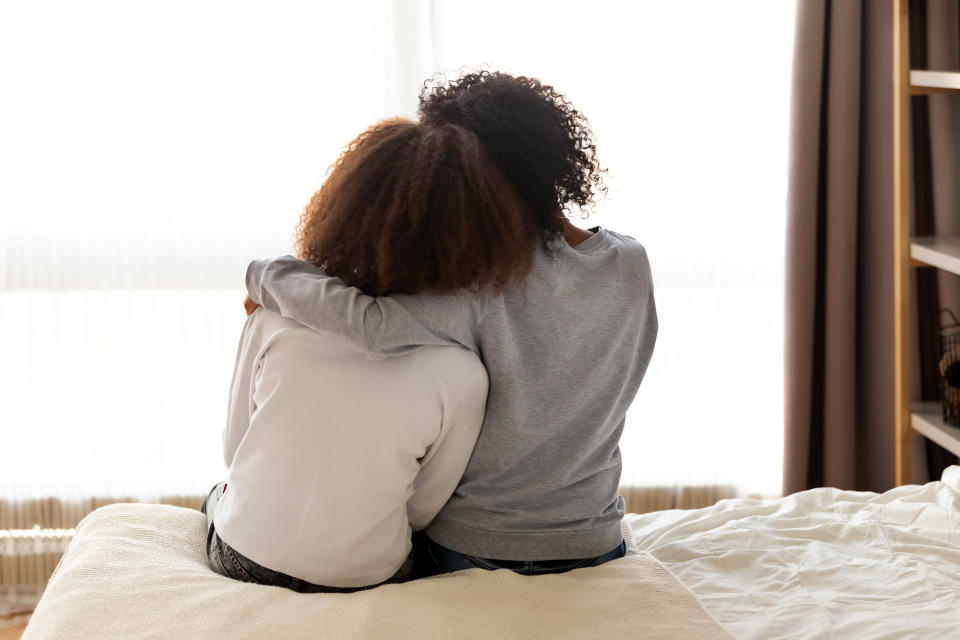 Rear back view black mother and daughter embrace sitting on bed at home