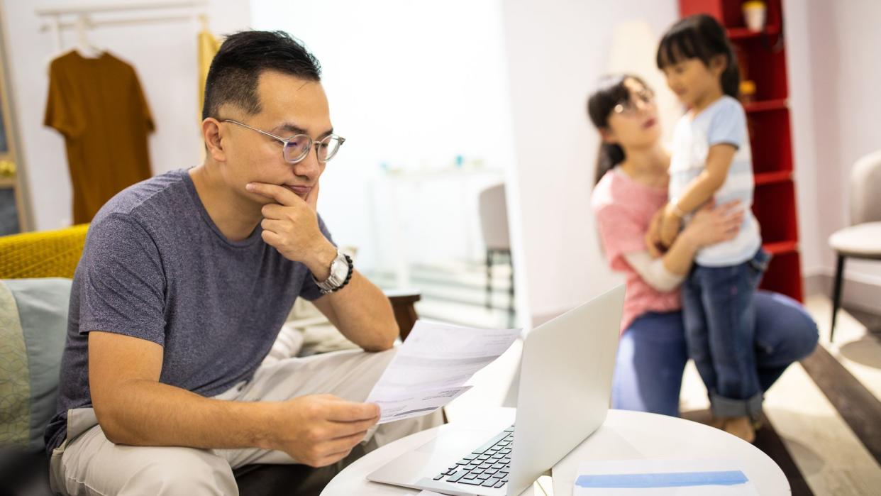 Worried man sitting on sofa in living room and holding paper, frustrated because of financial problems, wife and daughter playing near him.