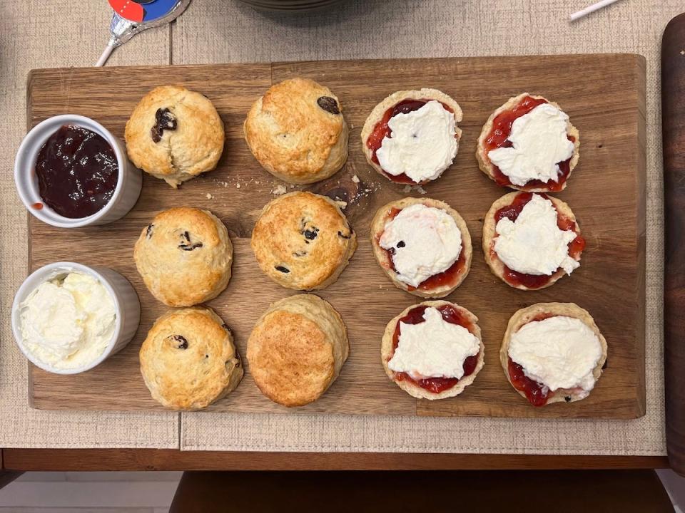 A wooden board with six whole scones, and six halves of scones covered with cream and jam, next to white ramekins containing strawberry jam and a faux clotted cream.