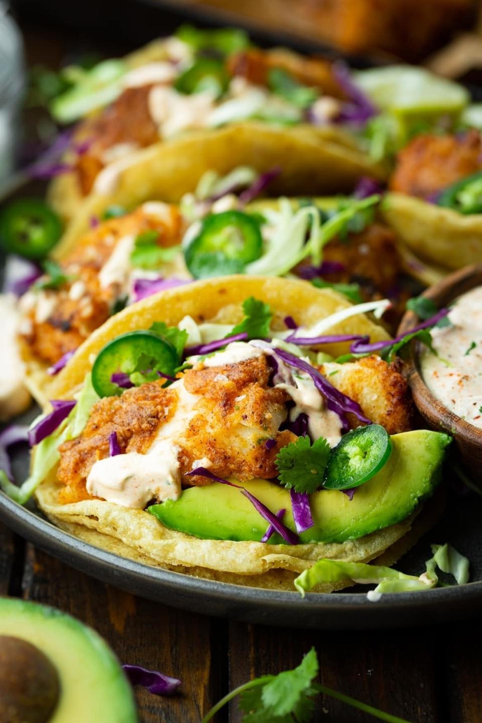 Plate of tacos filled with crispy fish, avocado slices, shredded cabbage, jalapeno slices, cilantro, and a creamy sauce