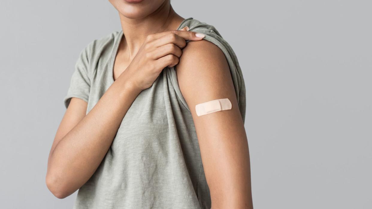  photo shows a young woman with light brown skin lifting up the sleeve of her t shirt to show a bandage on her arm, implying she recently got an injection 