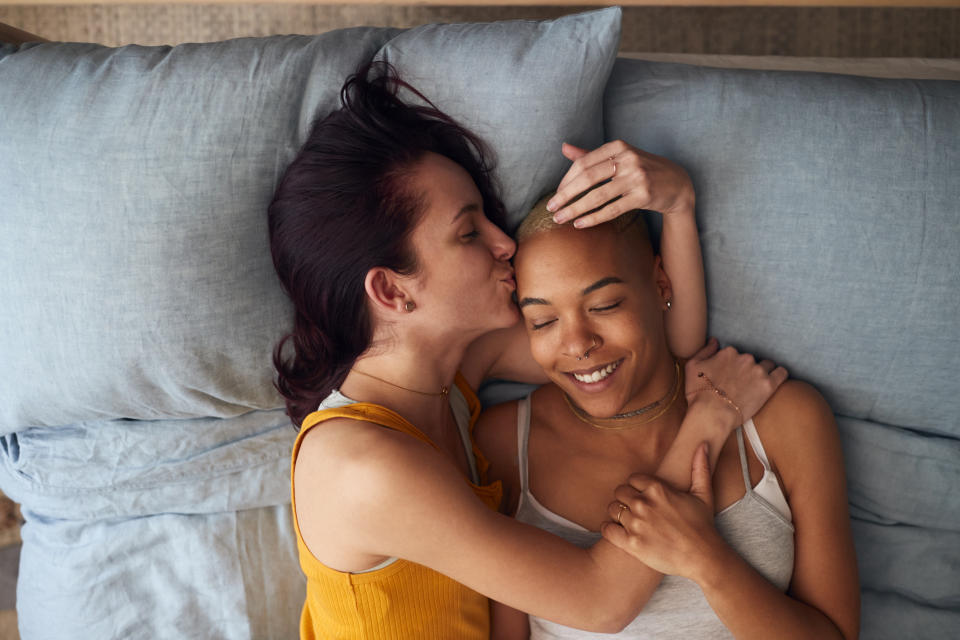 Two people lie on a bed. One with a shaved head smiles as the other with longer hair kisses their forehead. Both look content and relaxed