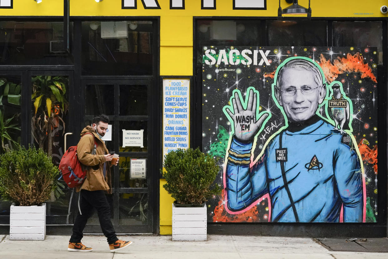A pedestrian wears his mask below on his chin as he walks past a street art depiction on Dr. Anthony Fauci, Friday, March 26, 2021, in the East Village neighborhood of New York. A year after becoming a global epicenter of the coronavirus pandemic, New York and New Jersey are back atop the list of U.S. states with the highest rates of infection. (AP Photo/Mary Altaffer)