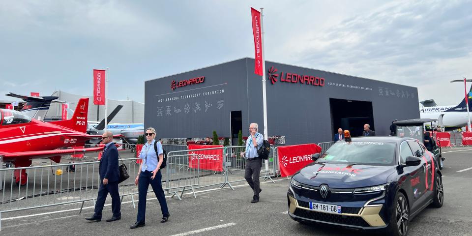 People walk past a small grey temporary building with the Leonardo logo on, and a red plane next to it, at the paris air show.
