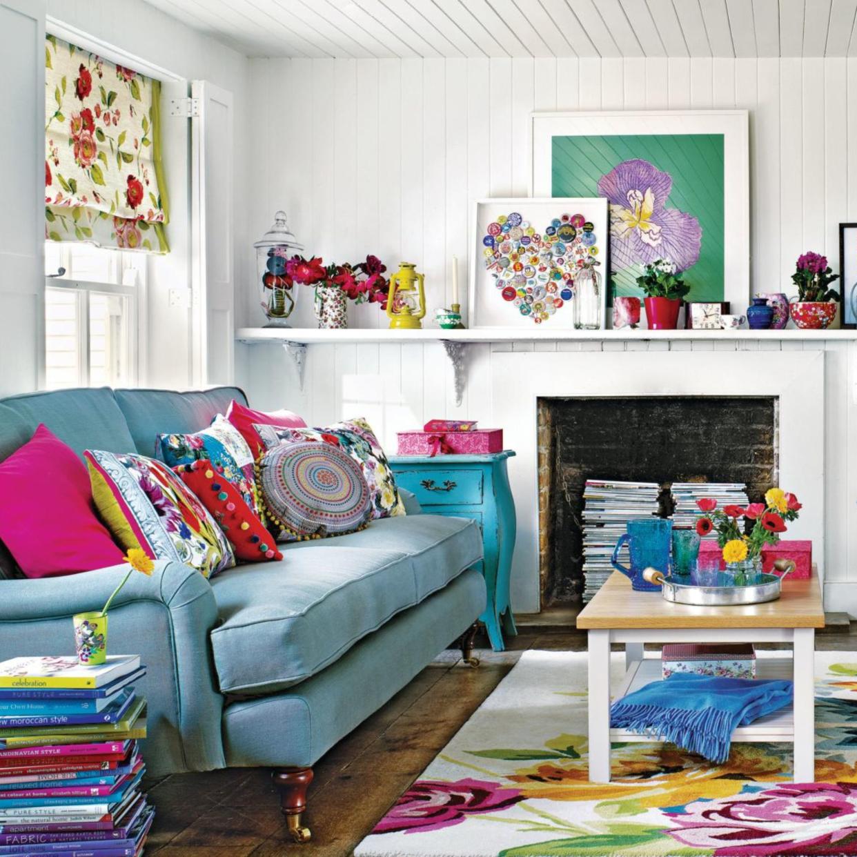  White living room, blue sofa with bright multicoloured cushions, bold floral rug, wooden coffee table, white sofa, stacked books used as table, stacked magazines in fireplace, wooden floorboards, 3D heart art on shelf, potted house plants. 